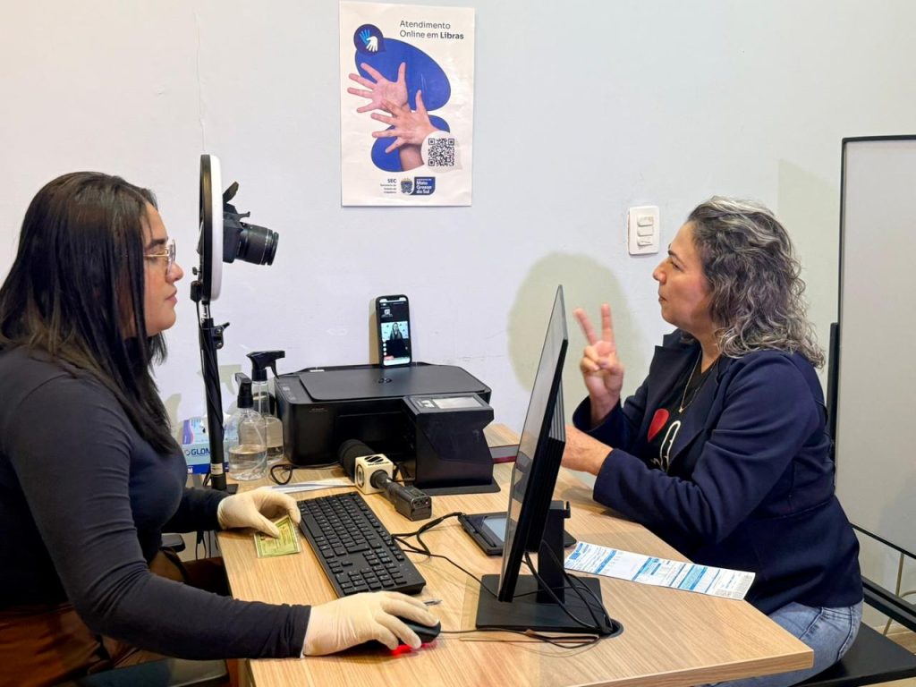 Duas mulheres sentadas frente a frente em uma mesa de escritório. À esquerda, uma mulher usa luvas e opera um computador. À direita, outra mulher gesticula em Libras (Língua Brasileira de Sinais). Sobre a mesa, há um monitor, impressora e um tripé com iluminador de LED (ring light).