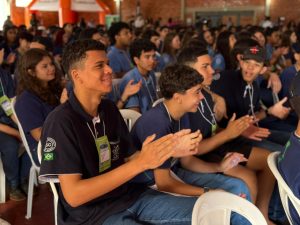 Um grande grupo de jovens está sentado em cadeiras brancas em um auditório ou ginásio. Em destaque, no primeiro plano, um jovem sorri e aplaude, vestindo uma camiseta polo azul-marinho com o brasão de São Gabriel do Oeste e a bandeira do Brasil na manga. Ao fundo, dezenas de outros estudantes com uniformes semelhantes participam do evento.
