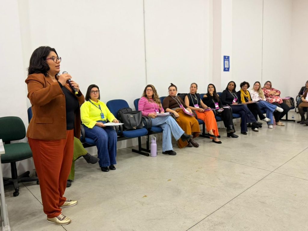 Foto em plano médio de uma sala de conferências. À esquerda, uma mulher de pele morena, óculos de grau e cabelos escuros curtos fala ao microfone. Ela usa um blazer marrom e calça terracota. À direita, enfileiradas em cadeiras azuis encostadas na parede branca, estão outras nove mulheres sentadas, observando e ouvindo atentamente. Elas usam roupas coloridas e casuais.