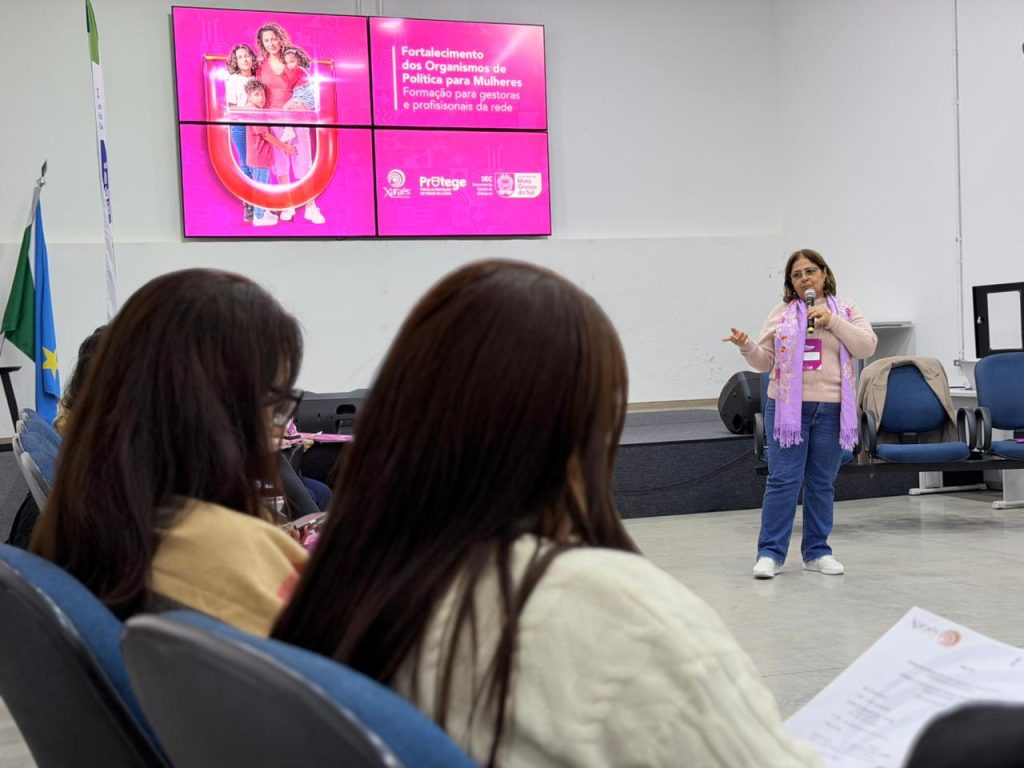 Vista em perspectiva de um auditório, mostrando uma mulher de pele clara, óculos e blusa rosa com lenço lilás falando ao microfone em cima do palco. Ao fundo, um grande telão rosa exibe o texto "Fortalecimento dos Organismos de Política para Mulheres". Em primeiro plano, parte da plateia assiste ao evento de costas para a câmera, com foco nas cabeças e ombros de duas mulheres sentadas.