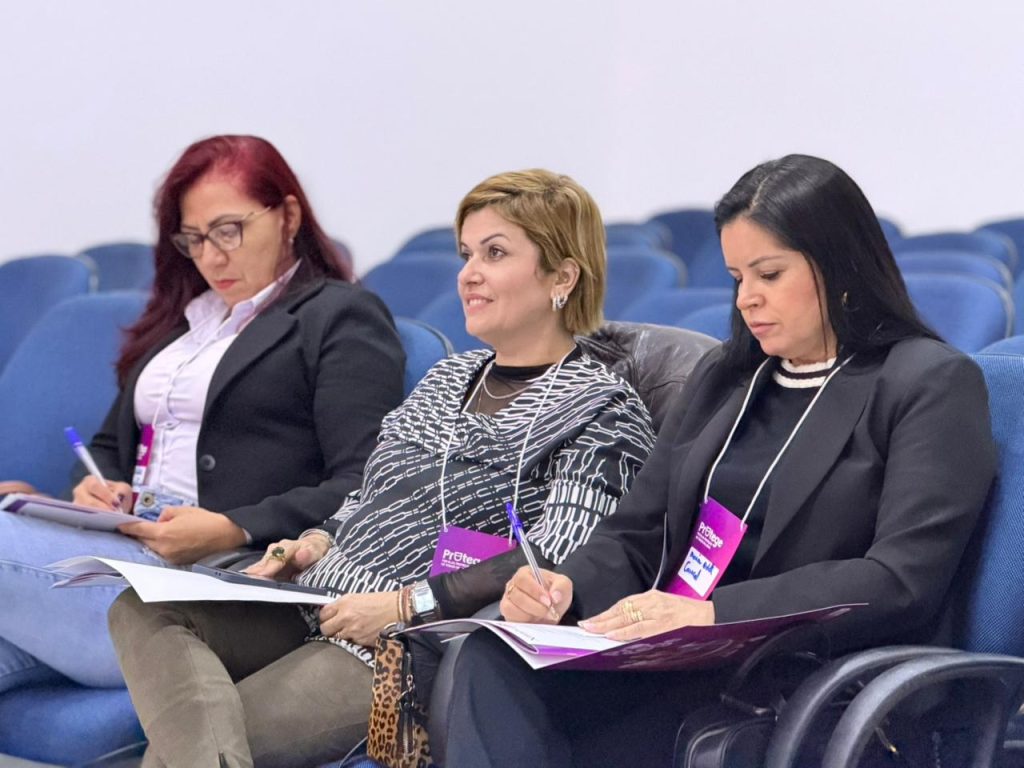 Três mulheres sentadas lado a lado nas cadeiras azuis do auditório, com crachás rosa pendurados no pescoço. A mulher do meio sorri olhando para a frente, enquanto as das pontas fazem anotações em cadernos sobre o colo.