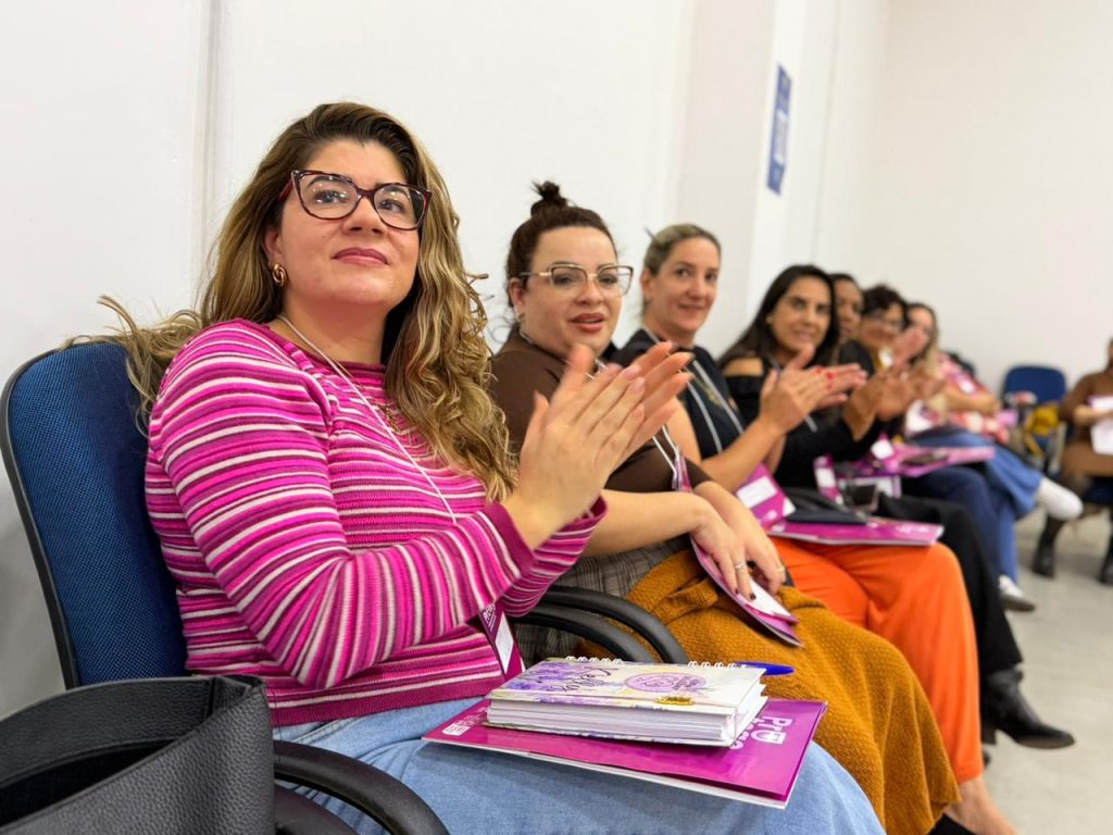 Foto em plano médio e fechado, em ângulo diagonal, mostrando um grupo de mulheres sentadas em cadeiras azuis. Em primeiro plano e em destaque, uma mulher de óculos de armação escura, cabelos longos ondulados com luzes e blusa listrada em tons de rosa e branco aparece aplaudindo com um sorriso discreto. No seu colo há um caderno e uma pasta roxa. Ao fundo, outras mulheres na fileira também são vistas aplaudindo, com o foco diminuindo gradativamente ao longo da linha.