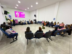 Foto em plano geral de uma sala ampla com piso de cimento queimado claro e paredes brancas. Cerca de vinte e cinco mulheres e um homem estão sentados em cadeiras estofadas azuis, dispostas em um grande semicírculo. No centro do círculo, uma mulher agachada fotografa o evento. Ao fundo, uma tela digital dupla exibe um slide rosa com o texto "Fortalecimento dos Organismos de Políticas para Mulheres". No canto esquerdo, há uma bandeira do Brasil e um banner com o brasão do Governo de Mato Grosso do Sul.