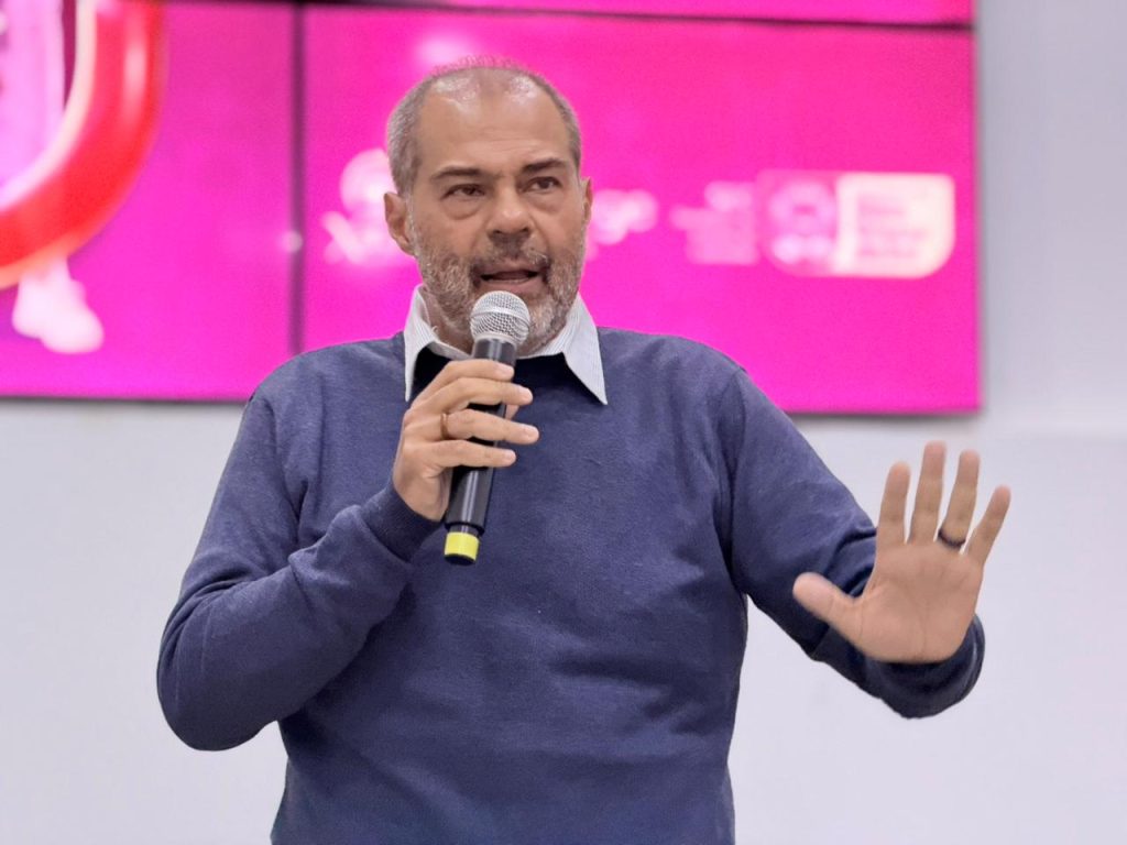 Homem de pele parda clara, cabelos curtos grisalhos e barba por fazer, vestindo blusa azul sobre camisa de colarinho claro. Ele fala ao microfone e faz um gesto com a mão esquerda aberta. Ao fundo, uma tela rosa vibrante com logotipos institucionais.