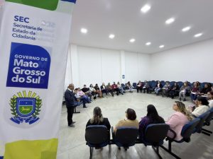 Amplo auditório com cadeiras azuis dispostas em formato de semicírculo, ocupadas por várias mulheres. À esquerda, o mesmo homem da imagem anterior fala em pé. Em primeiro plano, destaca-se um banner branco com o brasão e a inscrição "Governo de Mato Grosso do Sul".