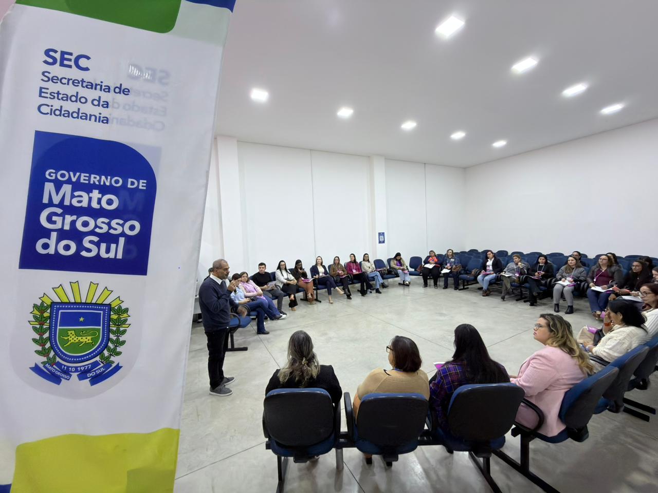 Amplo auditório com cadeiras azuis dispostas em formato de semicírculo, ocupadas por várias mulheres. À esquerda, o mesmo homem da imagem anterior fala em pé. Em primeiro plano, destaca-se um banner branco com o brasão e a inscrição "Governo de Mato Grosso do Sul".