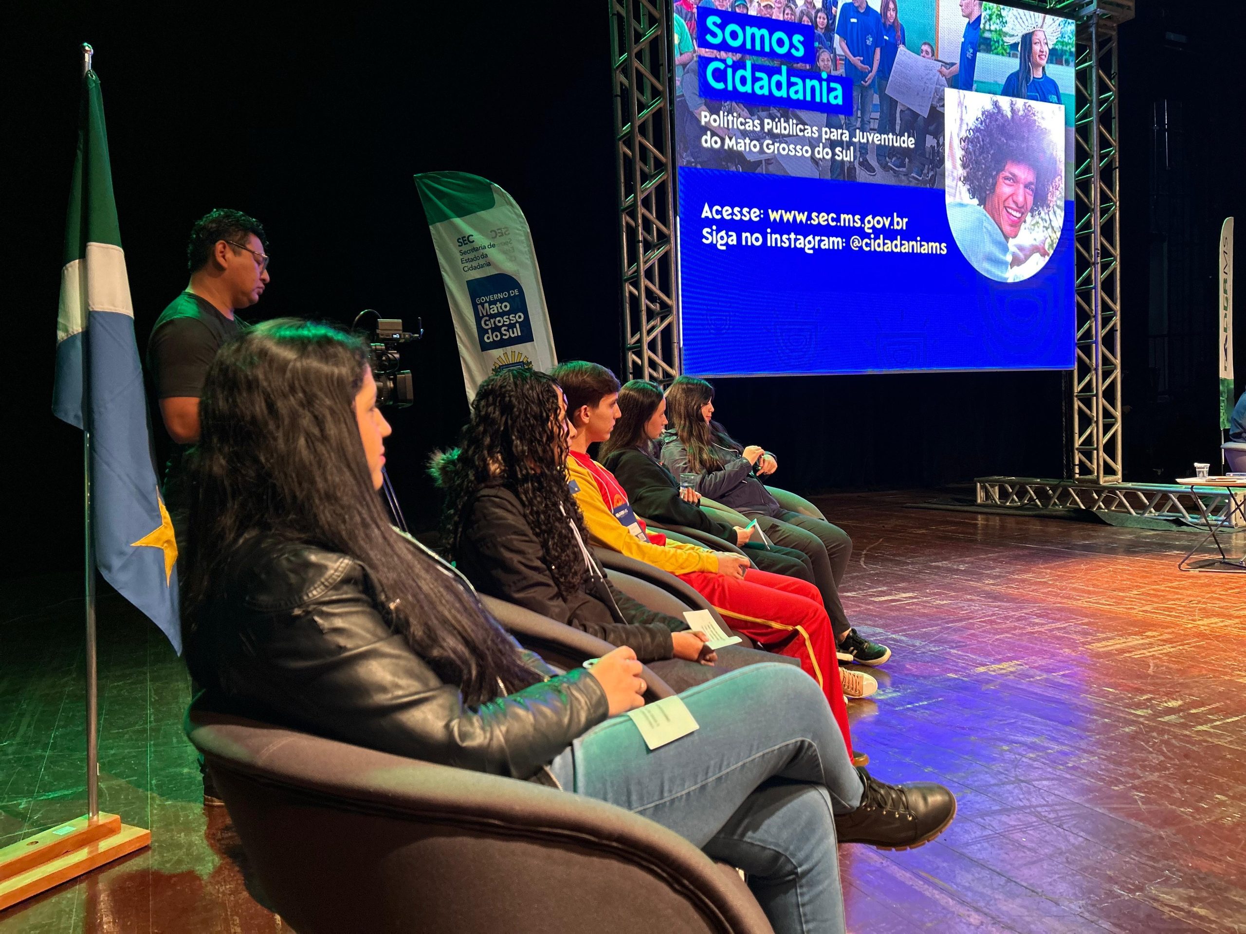 Jovens sentados em poltronas em um palco durante um evento. Eles estão de perfil, olhando para o centro do palco. Ao fundo, um telão exibe a frase "Somos Cidadania" e informações sobre políticas públicas para a juventude do Mato Grosso do Sul. À esquerda, vê-se a bandeira do estado.