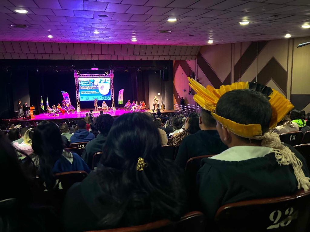 Vista ampla de um auditório. No palco iluminado, dois grupos de jovens estão sentados em semicírculo, voltados para um mediador ao centro. No fundo, um grande telão exibe a campanha "Somos Cidadania". Em primeiro plano, vê-se a silhueta da plateia sentada e um jovem de cocar amarelo.