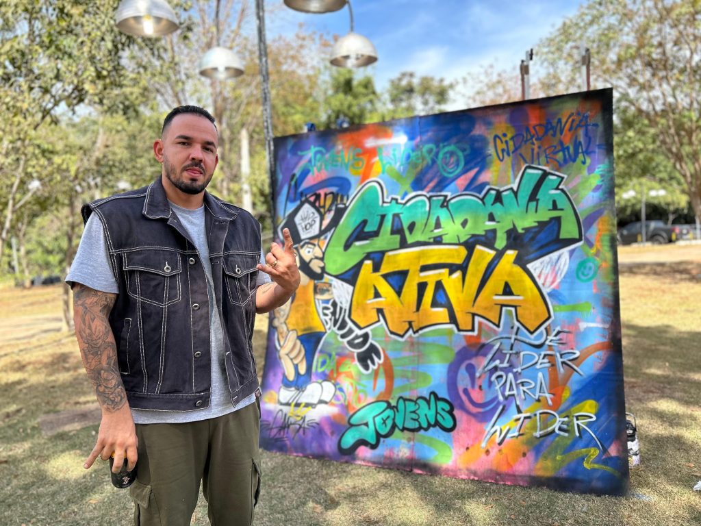 Um homem jovem em pé, ao ar livre, segurando uma lata de spray. Ao lado dele, um grande painel de grafite colorido com as palavras "Cidadania Ativa" em destaque. O cenário ao fundo possui árvores e um céu azul com nuvens leves.