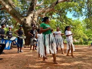 Fotografia colorida ao ar livre em um terreno de terra batida sob a copa de uma grande árvore. À esquerda, quatro homens tocam instrumentos de percussão (tambores e caixa). Ao centro e à direita, três mulheres e duas crianças realizam uma dança cultural. Elas vestem saias de fitas brancas sobre suas roupas e dançam descalças. O ambiente é ensolarado e cercado por vegetação verde.