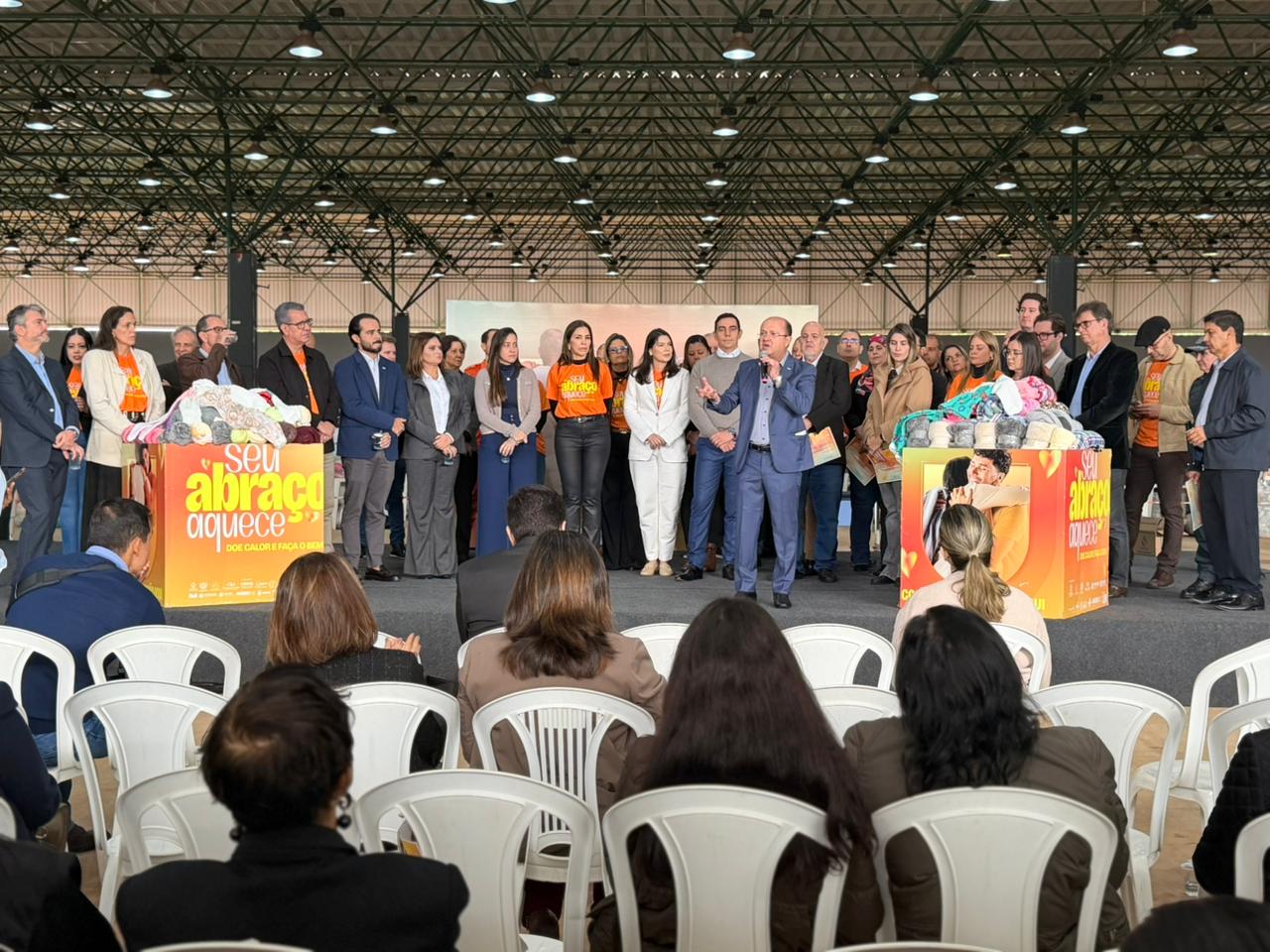 Fotografia em plano geral do mesmo palco no pavilhão. Agora, um homem de terno azul fala ao microfone no centro do palco, gesticulando com as mãos. O grupo de pessoas ao redor o escuta atentamente. As duas caixas amarelas com a campanha "Seu Abraço Aquece" delimitam as laterais do palco, repletas de donativos. Na plateia em primeiro plano, pessoas aparecem sentadas de costas.