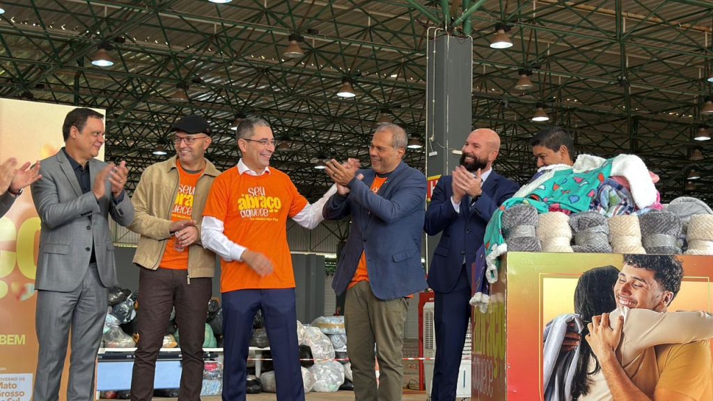 Fotografia em ambiente interno amplo onde seis homens, alinhados lado a lado, sorriem e aplaudem. Três deles vestem camisetas laranjas com a frase "seu abraço aquece", enquanto os outros usam ternos e jaqueta. À direita, há uma grande caixa cheia de cobertores e mantas dobradas, decorada com a ilustração de um abraço na parte frontal. Ao fundo, nota-se a estrutura metálica do teto de um galpão.
