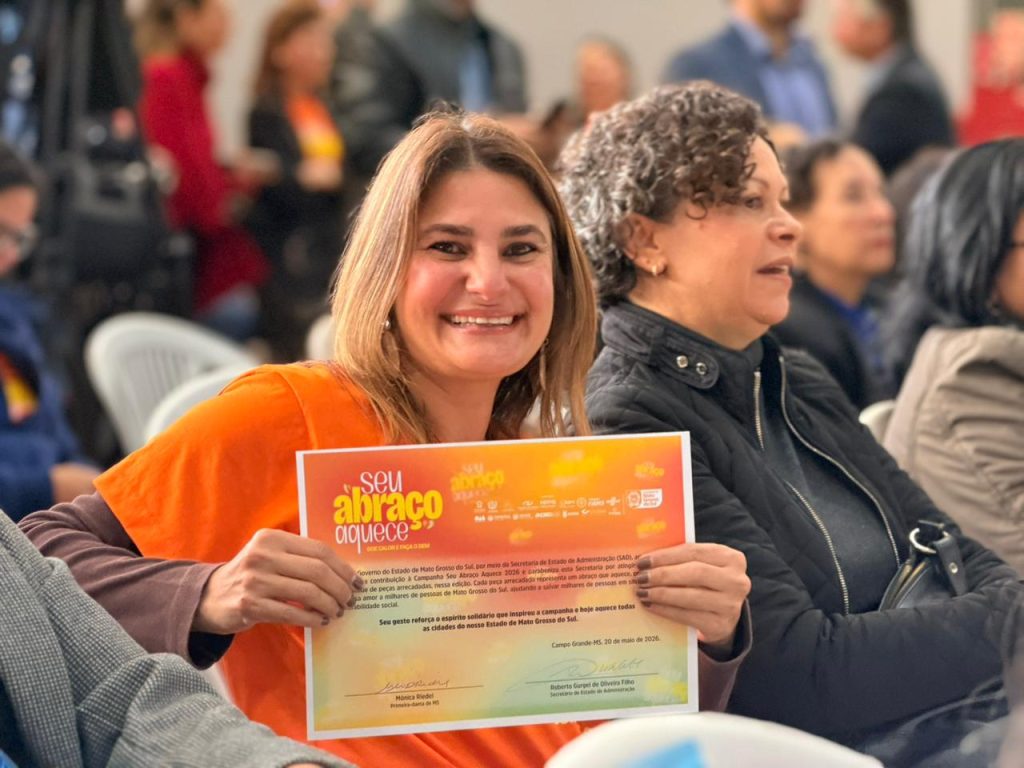 Fotografia em plano médio focado em uma mulher loira na plateia que sorri para a câmera. Ela segura com as duas mãos um certificado oficial da campanha "Seu Abraço Aquece", assinado pela Primeira-dama Mônica Riedel e datado de 20 de maio de 2026. Ela veste uma blusa laranja e está sentada ao lado de outra mulher em um ambiente com público ao fundo.