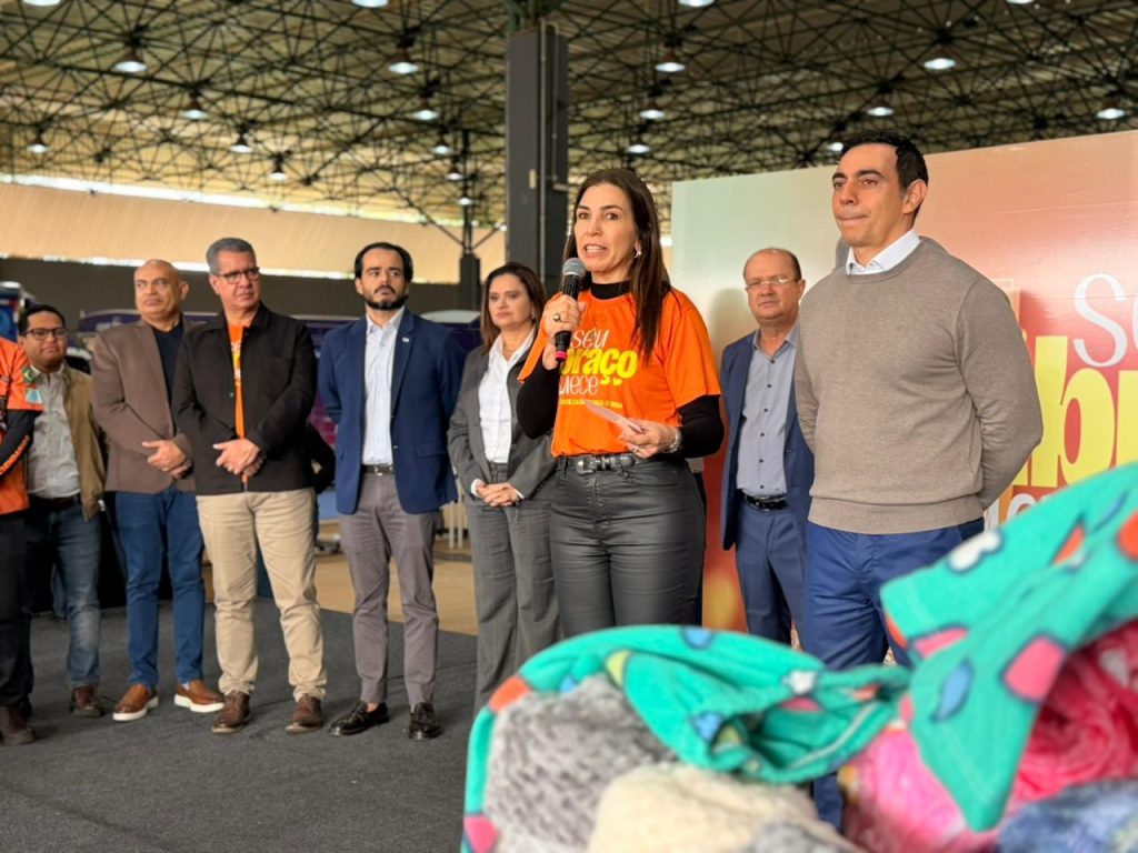 Fotografia em plano médio de uma mulher de cabelos castanhos e compridos falando ao microfone em um palco. Ela veste uma camiseta laranja com a inscrição "Seu Abraço Aquece" e calça preta justa. Ao seu lado e ao fundo, um grupo de homens e mulheres em trajes sociais e casuais acompanha o discurso. Em primeiro plano à direita, aparecem cobertores dobrados em tons de verde e rosa.