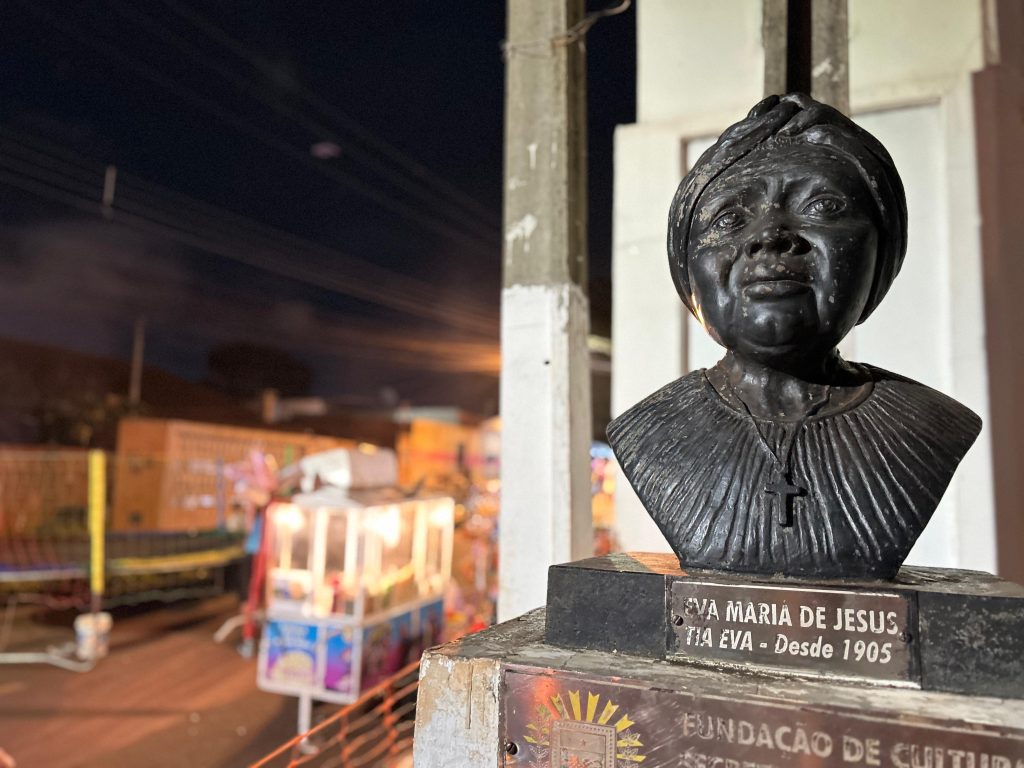 Fotografia noturna em plano médio de um busto de bronze representando Eva Maria de Jesus (Tia Eva). A escultura retrata uma mulher negra com um turbante e um colar de crucifixo. Abaixo, uma placa de identificação diz "Eva Maria de Jesus - Tia Eva - Desde 1905". Ao fundo, de forma desfocada, vê-se a iluminação de uma festa popular com barracas e um pula-pula sob o céu noturno.