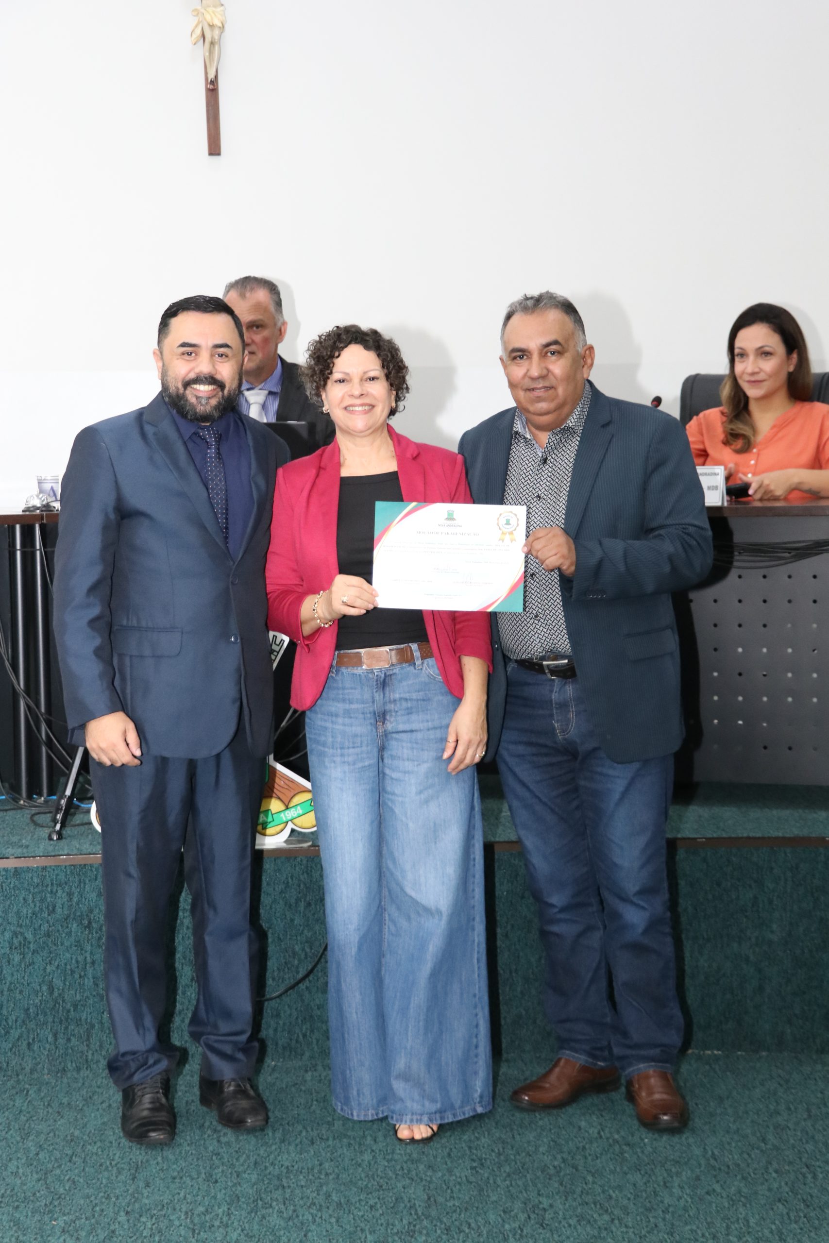 Três pessoas posam sorrindo em um plenário. À esquerda, um homem de terno azul-marinho; ao centro, uma mulher de jaqueta rosa e calça jeans segurando um certificado; à direita, um homem de paletó azul e calça jeans. Ao fundo, outras pessoas aparecem sentadas na bancada.