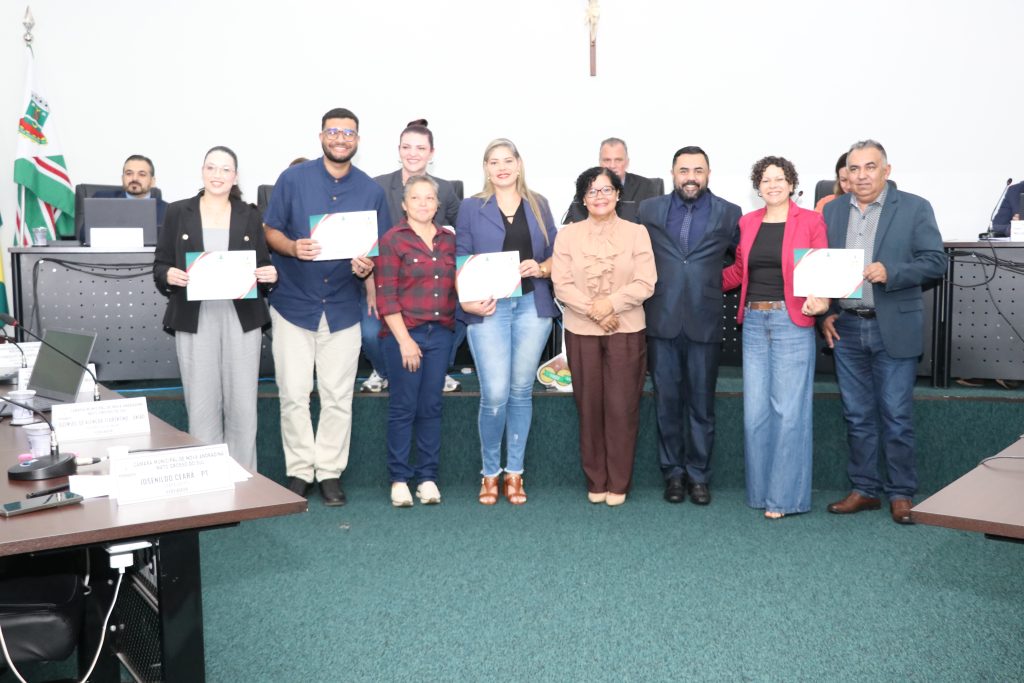 Foto em grupo no plenário da Câmara. Sete pessoas estão em pé, lado a lado, sorrindo para a câmera, e quatro delas seguram certificados de homenagem. Ao fundo, vereadores estão posicionados atrás de suas bancadas. O ambiente tem paredes brancas e carpete verde.