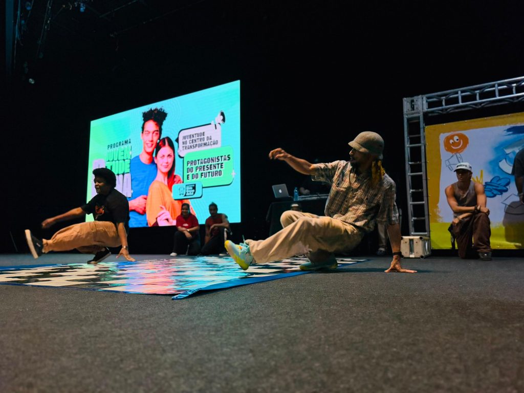Dois jovens realizam passos de breakdance em cima de um tapete quadriculado no palco de um auditório escuro. Ao fundo, um telão digital verde exibe a identidade visual do evento com os dizeres "Juventude no centro da transformação". Outros jovens sentados ao lado observam a apresentação.