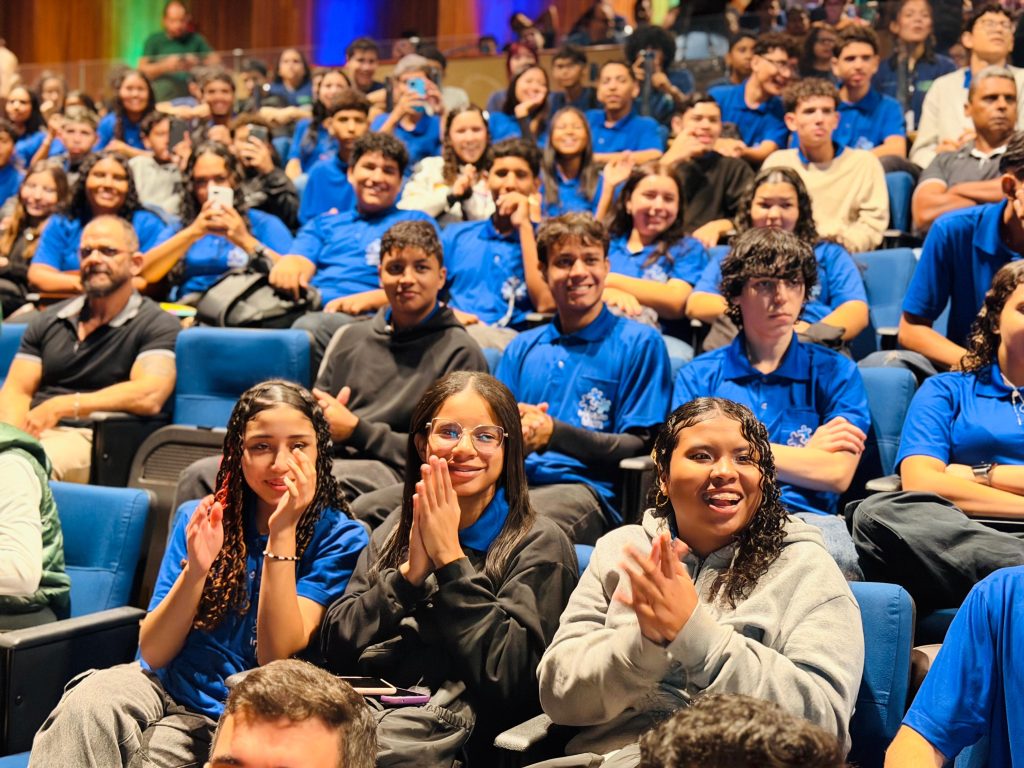 Em primeiro plano, três jovens sorriem e batem palmas sentadas nas poltronas de um auditório. A jovem do meio usa óculos de grau. Ao fundo, o restante da plateia, composta majoritariamente por jovens vestindo camisas polo azuis, acompanha o evento com atenção.