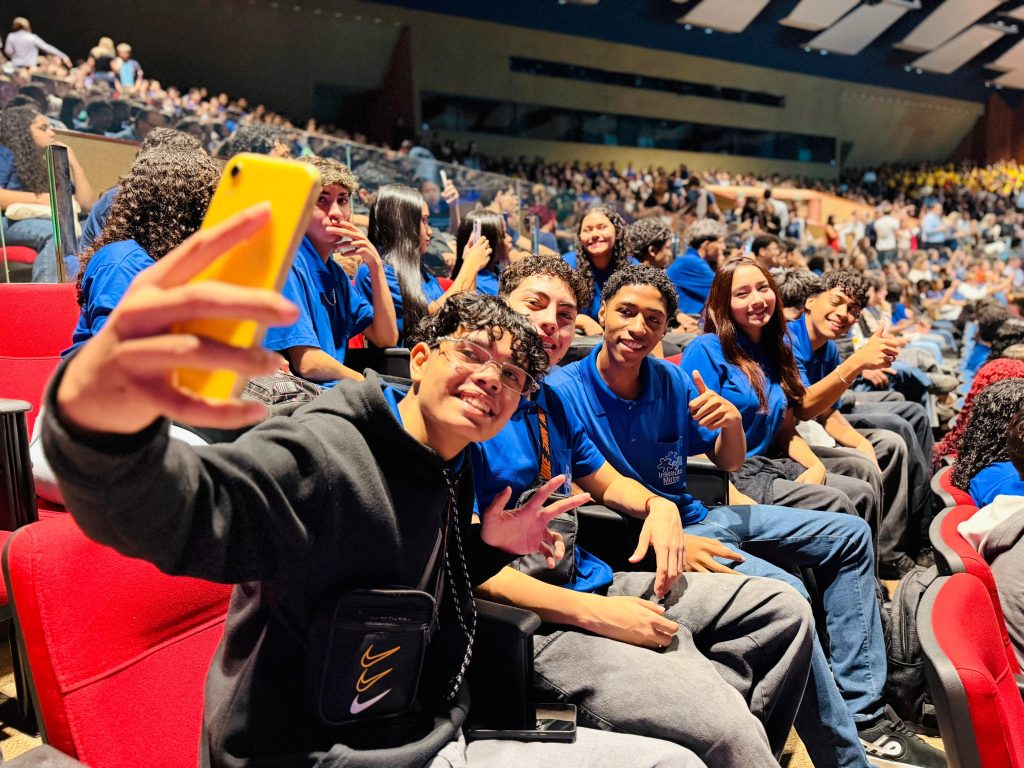 Um jovem em primeiro plano segura um celular com capinha amarela para tirar uma selfie com seus amigos nas poltronas vermelhas de um auditório. O grupo de jovens sorri e faz gestos de positivo e sinal de paz para a foto. Eles vestem camisetas azuis e pretas. Ao fundo, a arquibancada do auditório aparece repleta de pessoas.