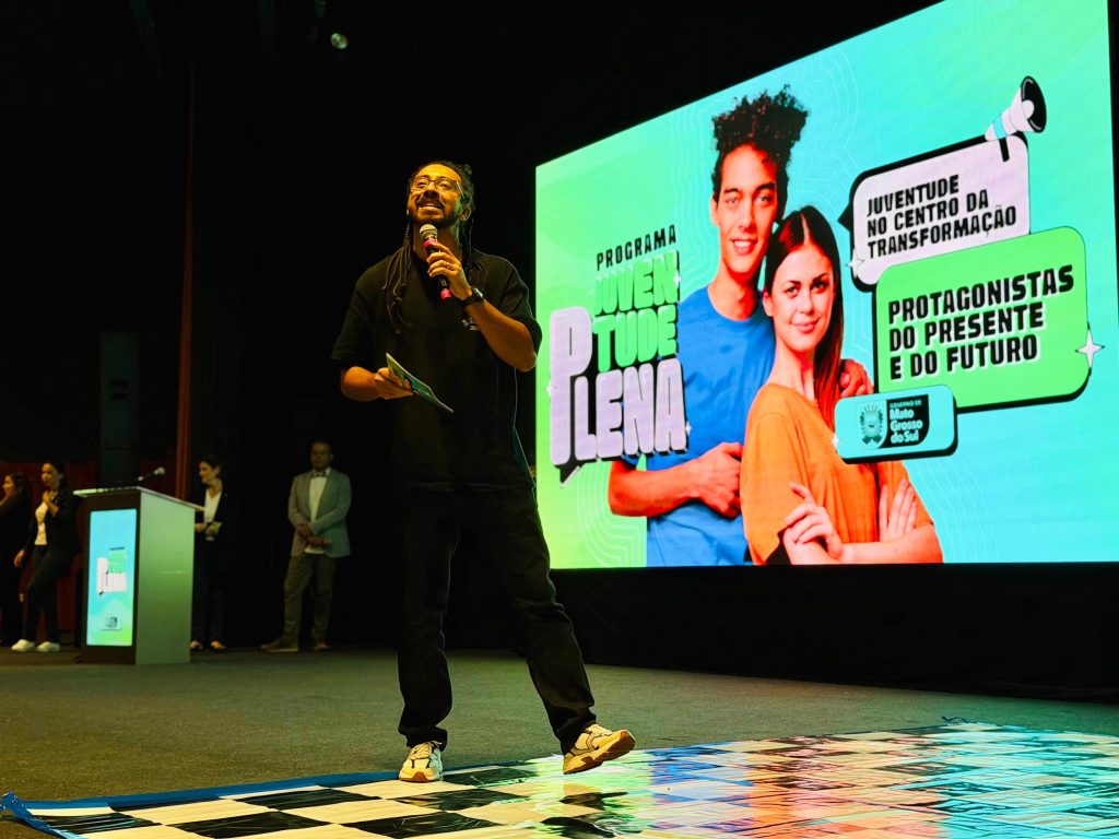 Um homem com dreadlocks, óculos e roupas escuras fala ao microfone no palco, sorrindo e olhando para cima. Atrás dele, há um enorme telão digital verde com a arte do evento "Programa Juventude Plena" e a frase "Protagonistas do presente e do futuro". O chão à sua frente tem uma lona quadriculada.