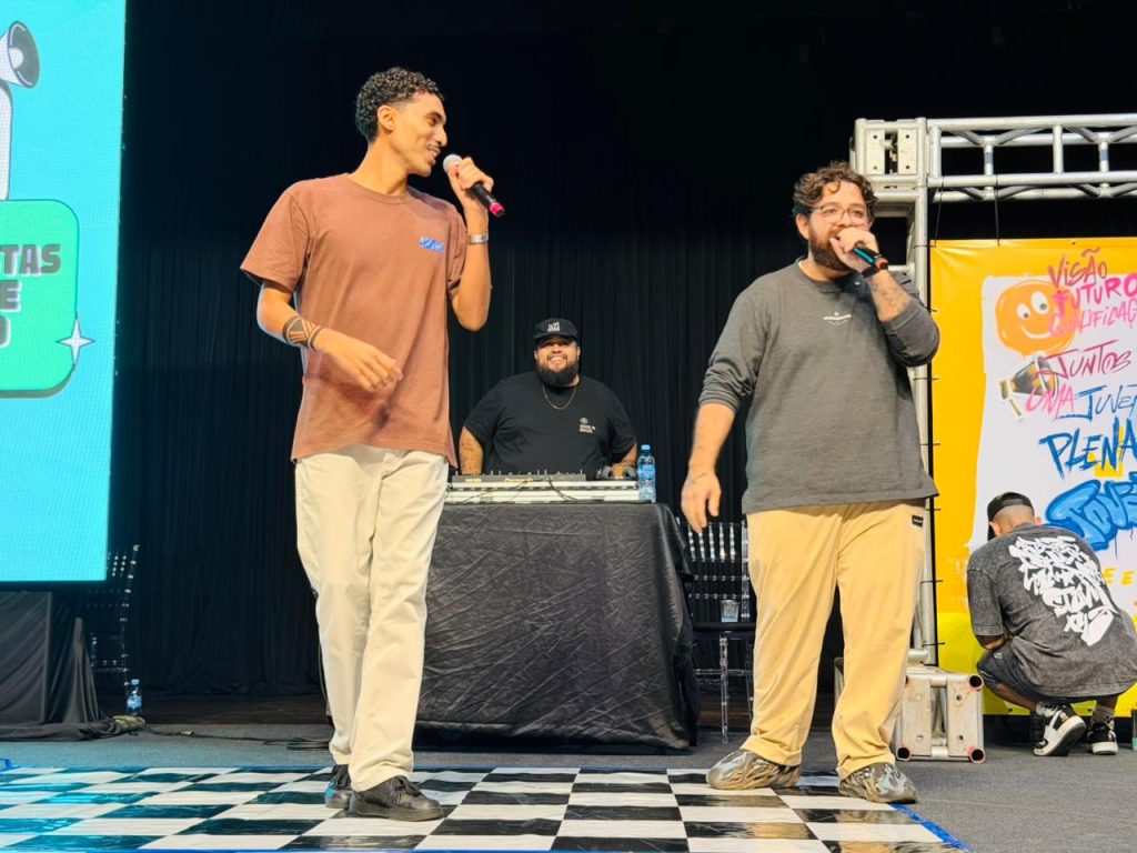 Dois jovens em cima de um palco sorriem e interagem usando microfones. O da esquerda veste camiseta marrom e calça clara, e o da direita usa óculos, blusa cinza e calça bege. Ao fundo, um DJ em sua mesa acompanha a apresentação diante de uma cortina preta. O chão do palco possui uma estampa quadriculada em preto e branco.