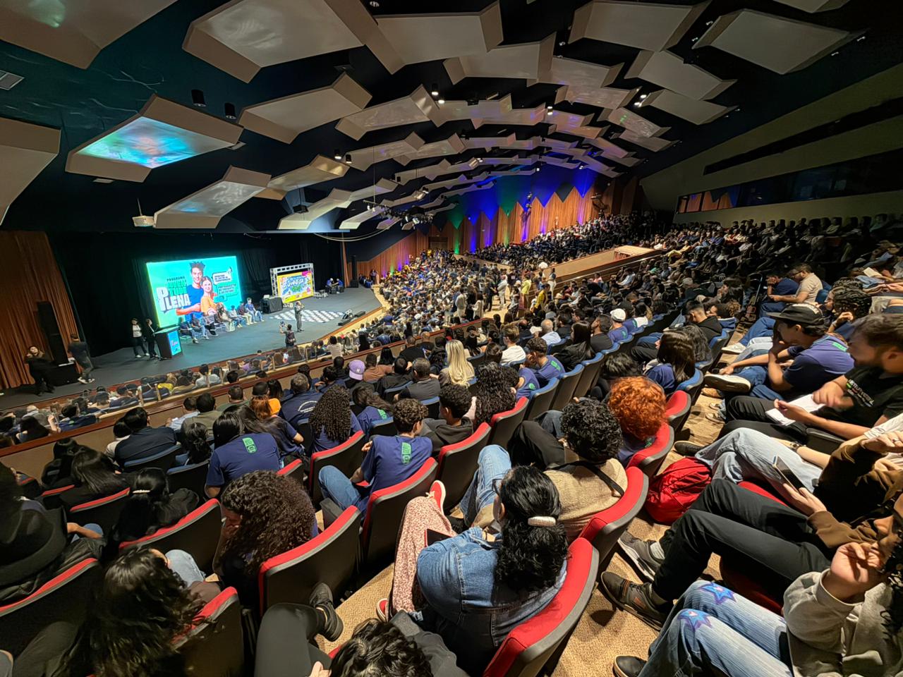 Foto em ângulo aberto e panorâmico mostrando a plateia lotada de um grande auditório. Centenas de pessoas estão sentadas em poltronas vermelhas e azuis dispostas em fileiras inclinadas. Ao fundo, o palco iluminado exibe um grande painel digital verde do "Programa Juventude Plena".