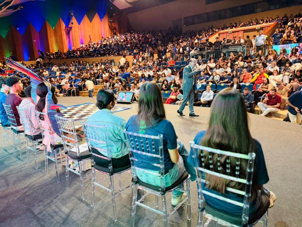 Um homem de terno cinza fala ao microfone no centro de um palco, de frente para um grande auditório lotado. Em primeiro plano, vistos de costas, estão jovens sentados em cadeiras transparentes; uma das jovens usa um cocar indígena com penas coloridas.