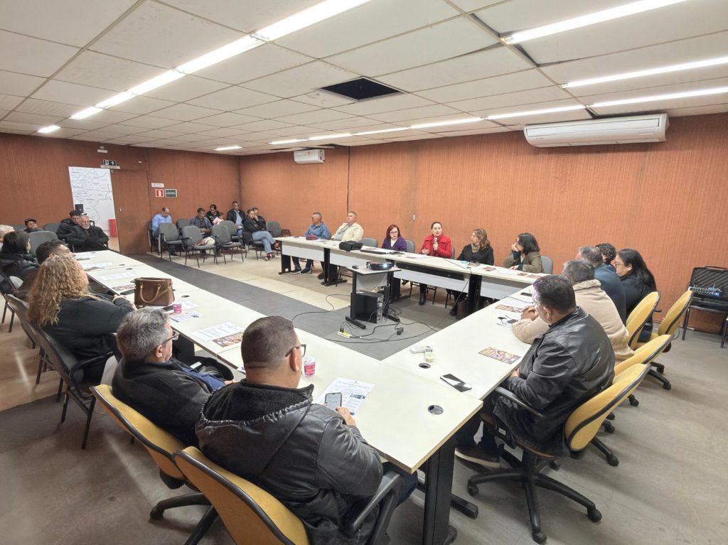 Fotografia de plano aberto em ângulo levemente elevado mostrando uma reunião em uma sala com paredes de madeira. Homens e mulheres estão sentados ao redor de mesas brancas dispostas em formato de "U". Ao fundo, uma mulher vestindo um casaco vermelho fala ao microfone. Há papéis e copos sobre as mesas.