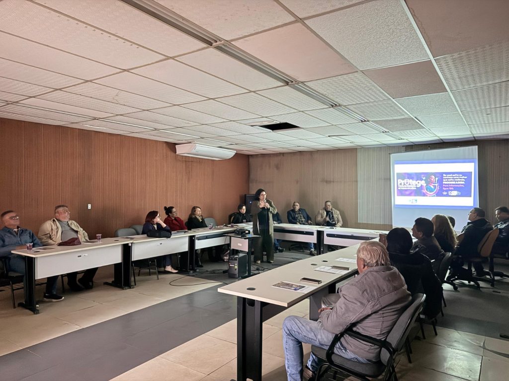 Vista panorâmica da sala de reunião. No centro, uma mulher de casaco verde oliva está de pé falando ao grupo. À direita, um projetor exibe em uma tela branca um slide da campanha "Protege", com tons de roxo e a imagem de uma mulher. Os participantes acompanham a apresentação sentados em mesas organizadas em semicírculo.