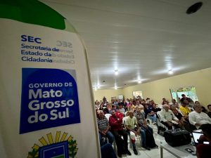 Foto do interior de um salão em Campo Grande (MS) repleto de pessoas sentadas em cadeiras brancas, acompanhando um evento. Em primeiro plano, à esquerda, destaca-se um banner com as logomarcas da Secretaria de Estado da Cidadania e do Governo de Mato Grosso do Sul. O ambiente é bem iluminado, com paredes claras e teto branco.