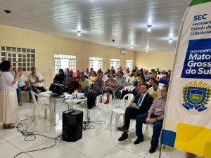 Fotografia horizontal em plano aberto de uma palestra em um auditório simples com piso de cerâmica branca. À esquerda, uma mulher em pé fala ao microfone para uma plateia de cerca de 50 pessoas, majoritariamente adultas e idosas, sentadas em cadeiras plásticas brancas. No centro, há uma mesa pequena com um projetor e equipamentos de som. No canto direito, em primeiro plano, aparece parte de um banner branco com os logotipos da "SEC - Secretaria de Estado da Cidadania" e do "Governo de Mato Grosso do Sul". O ambiente é iluminado por lâmpadas de LED no teto e luz natural vinda de janelas ao fundo.