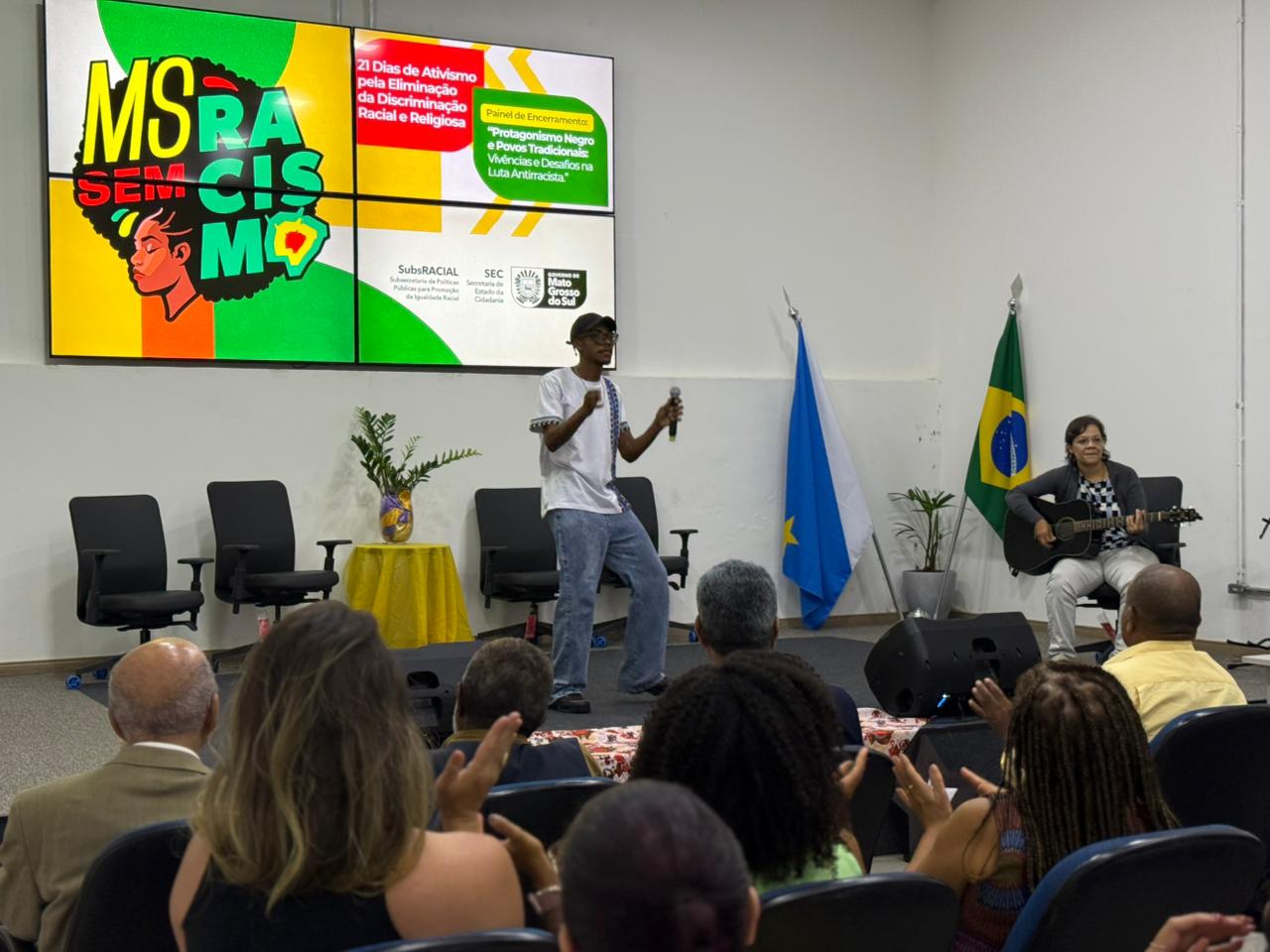Em um auditório, um jovem negro com boné e camiseta branca fala ao microfone no centro do palco. Ao lado dele, uma mulher sentada toca violão. Ao fundo, um painel digital exibe a frase "MS Sem Racismo" e informações sobre os "21 Dias de Ativismo pela Eliminação da Discriminação Racial e Religiosa". Parte da plateia é vista de costas em primeiro plano.
