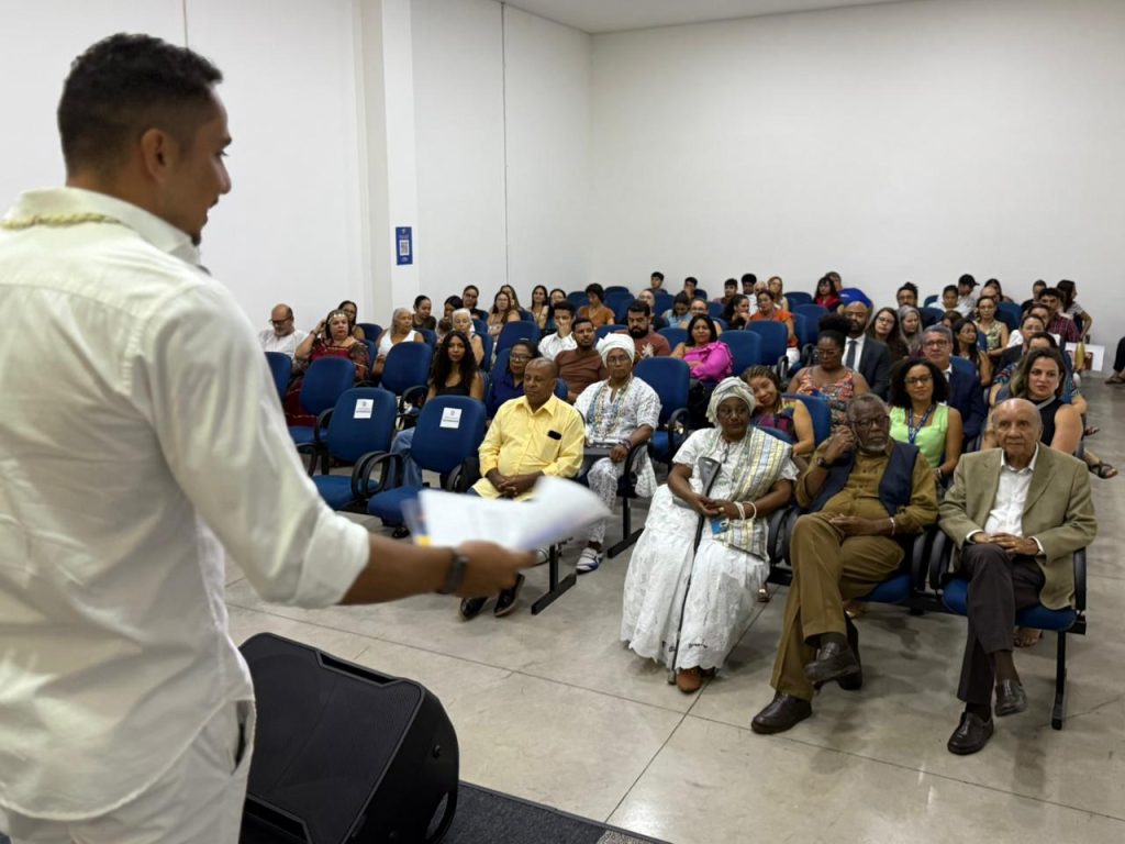Vista de cima do palco para a plateia de um auditório. Um homem de camisa branca está em primeiro plano, de costas, falando para o público. A plateia é diversa e está sentada em poltronas azuis; na primeira fila, destacam-se lideranças negras e religiosas vestidas com roupas tradicionais brancas e trajes formais.