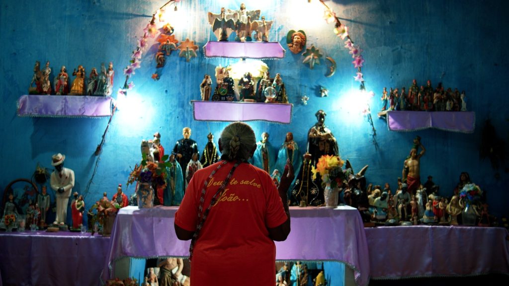 Uma mulher de costas, vestindo uma camiseta vermelha com a inscrição "Deus te salve João...", diante de um altar iluminado e repleto de diversas estátuas religiosas de santos e entidades, contra uma parede azul vibrante.