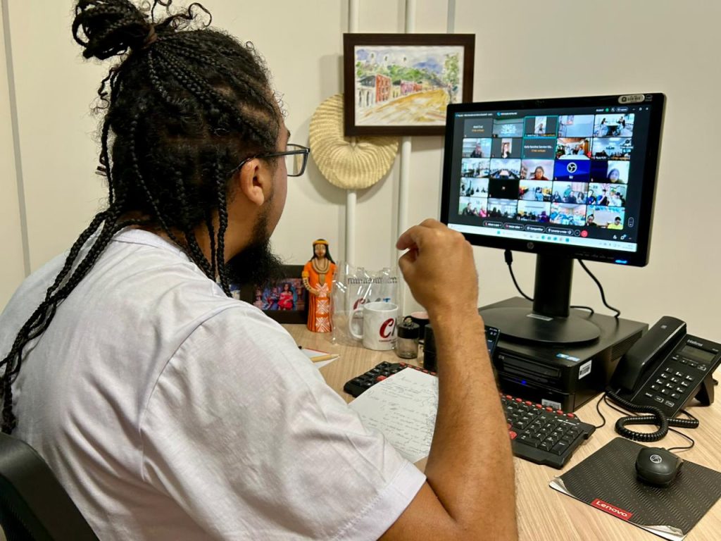 Vista lateral de um homem com cabelos longos em tranças (box braids) presos em um coque, usando camiseta branca e óculos. Ele está sentado em uma mesa de escritório, de frente para um monitor que exibe uma videoconferência com vários participantes. Sobre a mesa, há um teclado, papéis com anotações, um telefone e pequenos objetos decorativos. Ao fundo, uma parede branca com um quadro pequeno.