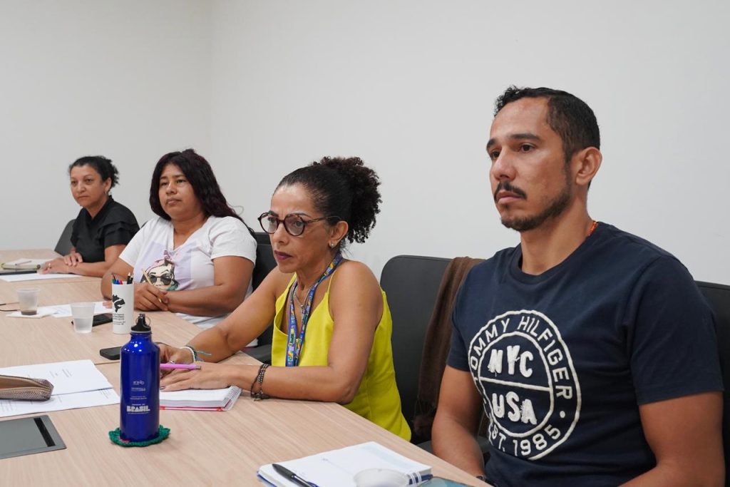Foto em plano médio e visão lateral de quatro pessoas sentadas enfileiradas ao longo de uma mesa de reunião clara. Em primeiro plano, à direita, está um homem pardo, de barba curta, vestindo camiseta preta com uma estampa circular escrita "Tommy Hilfiger". Ao lado dele, uma mulher negra de óculos, cabelos cacheados presos em um coque alto e blusa amarela, apoia as mãos sobre a mesa com um caderno aberto. Mais ao fundo, estão duas mulheres: uma de blusa branca com estampa e outra de blusa preta, ambas olhando atentamente na mesma direção, em um ambiente de escritório com paredes brancas.