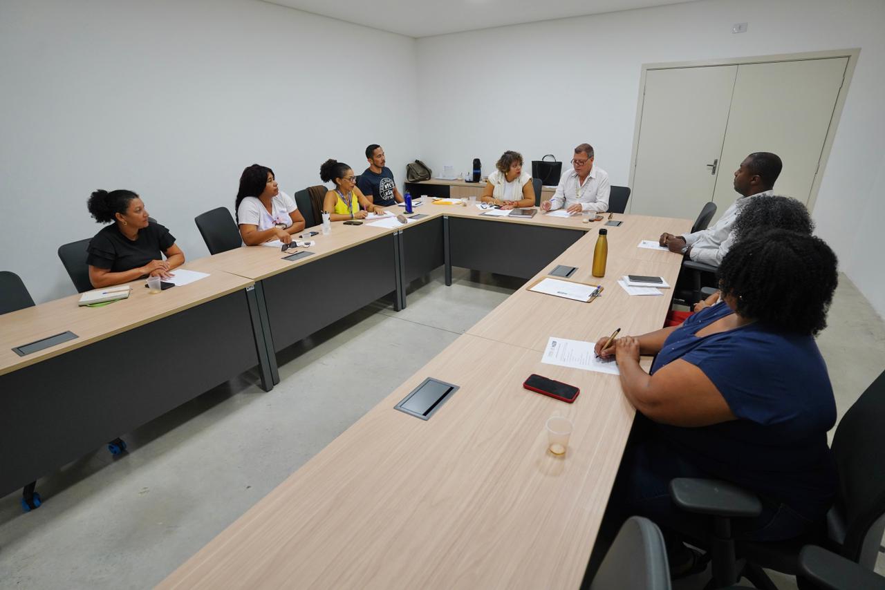 Foto em ângulo superior e plano aberto mostrando uma sala de reunião com paredes brancas. Nove pessoas estão distribuídas ao redor de mesas modulares dispostas em formato de "U". Em primeiro plano, à direita, destaca-se uma mulher negra de costas, com cabelos cacheados volumosos e blusa azul, anotando em um papel. Do outro lado da mesa, os participantes estão sentados em cadeiras de escritório escuras, prestando atenção à reunião. Sobre as mesas há cadernos, papéis, copos d'água, garrafas de metal e celulares.