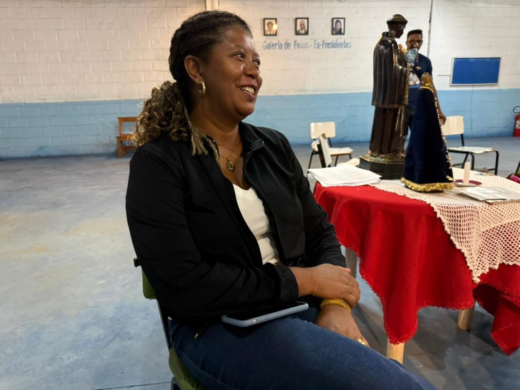 Mulher negra sorri de perfil, sentada em uma cadeira. Ela usa uma jaqueta preta sobre blusa branca e calça jeans. Ao fundo, há uma mesa com toalha vermelha e detalhes em crochê branco, onde estão posicionadas imagens religiosas, incluindo uma estátua de São Benedito e uma de Nossa Senhora Aparecida. A parede ao fundo exibe uma "Galeria de Fotos - Ex-Presidentes".