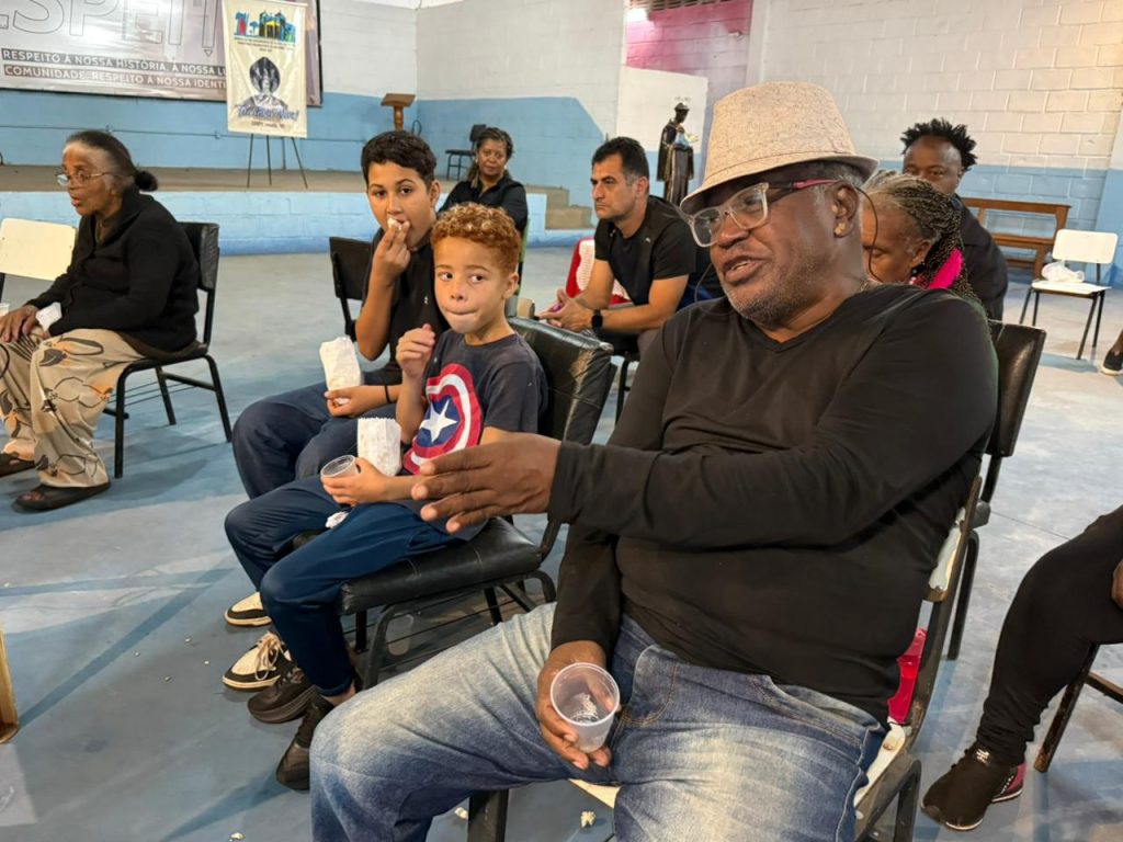 Um homem negro usando chapéu e óculos fala em primeiro plano, segurando um copo plástico. Ao lado dele, duas crianças comem pipoca, uma delas vestindo uma camiseta do Capitão América. Outras pessoas estão sentadas em cadeiras ao redor, assistindo e conversando no interior de um amplo galpão.