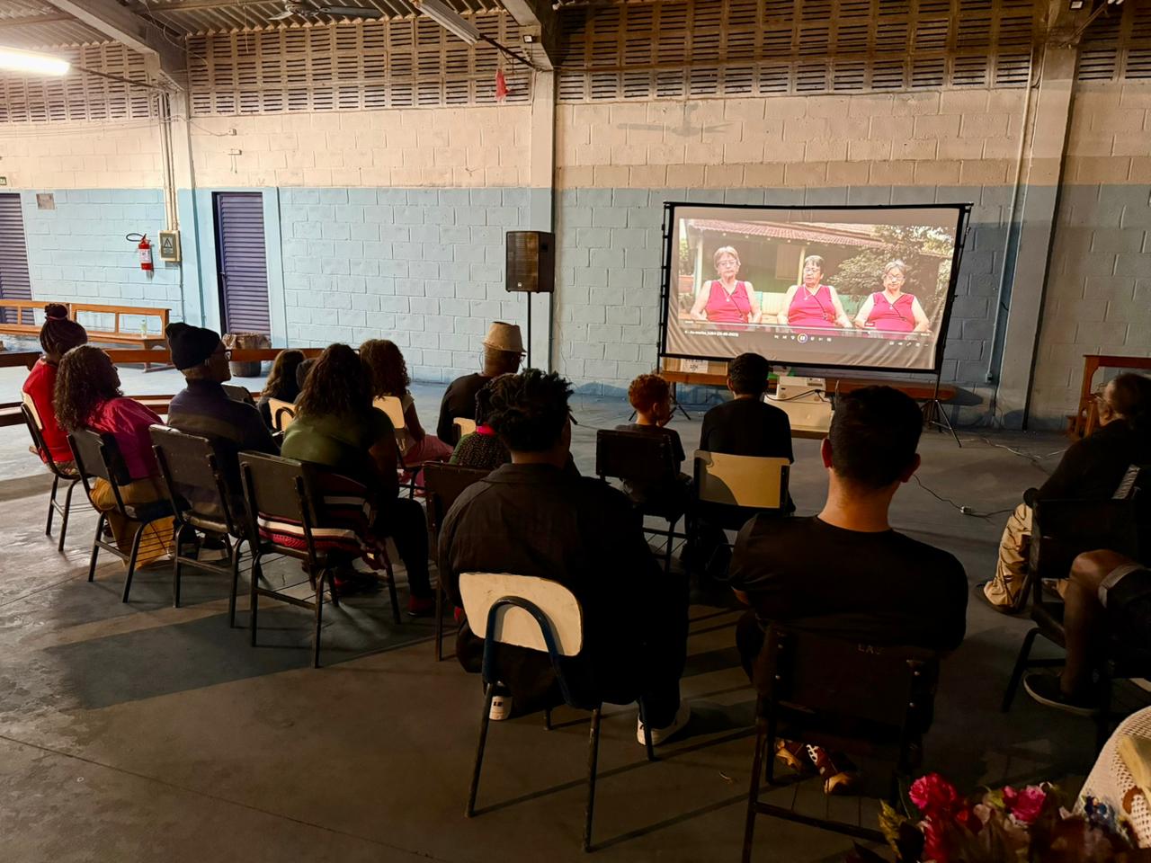 Público de costas assistindo a um filme em uma grande tela de projeção em um galpão amplo à noite. A tela exibe três mulheres idosas com blusas vermelhas idênticas, sentadas lado a lado. A iluminação do local é baixa, focada no telão.
