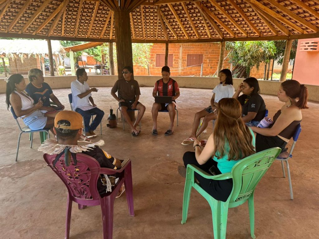 Fotografia em plano aberto mostrando um grupo de cerca de dez jovens indígenas sentados em círculo, em cadeiras de metal e plástico, dentro de uma grande estrutura circular com teto de palha (estilo oca ou maloca). Ao centro, um jovem segura um notebook. Os demais conversam e ouvem atentamente. Um dos jovens, em primeiro plano e de costas, usa um cocar de penas claras. O chão é de terra batida e, ao fundo, veem-se casas de alvenaria sob a luz do sol.