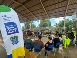 Fotografia em ângulo amplo de uma reunião realizada em uma quadra poliesportiva coberta. Dezenas de pessoas estão sentadas em círculo ocupando o espaço da quadra. Em destaque, no canto esquerdo do primeiro plano, há um banner vertical branco com os logotipos da "SEC - Secretaria de Estado da Cidadania" e do "Governo de Mato Grosso do Sul". Ao fundo, a estrutura da quadra é aberta, revelando árvores e o céu claro. À direita, uma mesa com toalha verde vibrante exibe vasos de cerâmica artesanal.