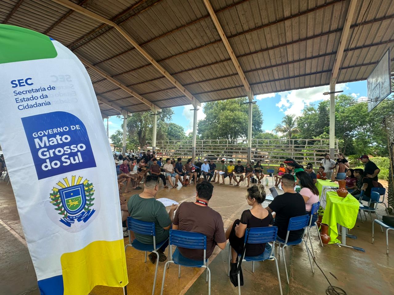 Fotografia em ângulo amplo de uma reunião realizada em uma quadra poliesportiva coberta. Dezenas de pessoas estão sentadas em círculo ocupando o espaço da quadra. Em destaque, no canto esquerdo do primeiro plano, há um banner vertical branco com os logotipos da "SEC - Secretaria de Estado da Cidadania" e do "Governo de Mato Grosso do Sul". Ao fundo, a estrutura da quadra é aberta, revelando árvores e o céu claro. À direita, uma mesa com toalha verde vibrante exibe vasos de cerâmica artesanal.