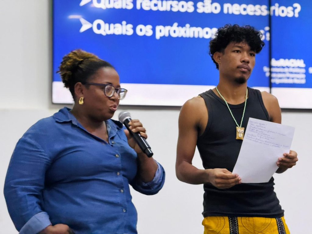 Uma mulher negra com óculos fala ao microfone ao lado de um jovem negro que segura uma folha de papel com anotações, durante uma apresentação no fórum.