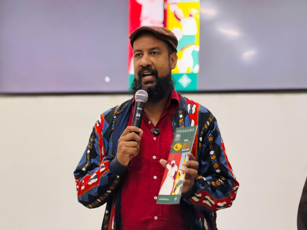 Um homem negro com barba e boina marrom fala ao microfone enquanto segura o Guia de Afroturismo. Ele veste uma jaqueta colorida com padrões geométricos.