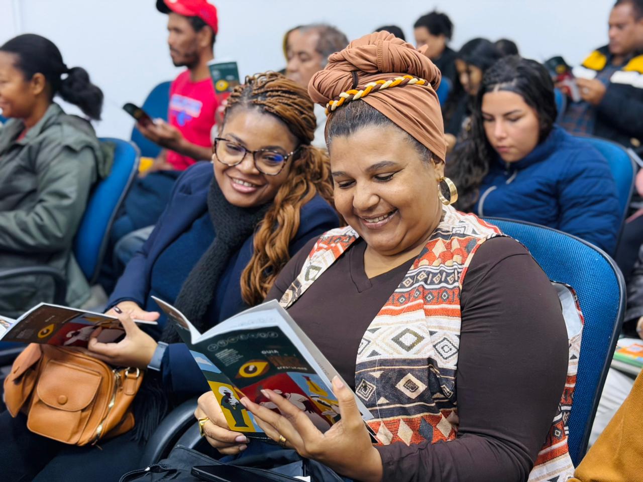 Duas mulheres negras sorridentes em uma plateia; a que está em primeiro plano usa um turbante marrom e lê atentamente o guia.