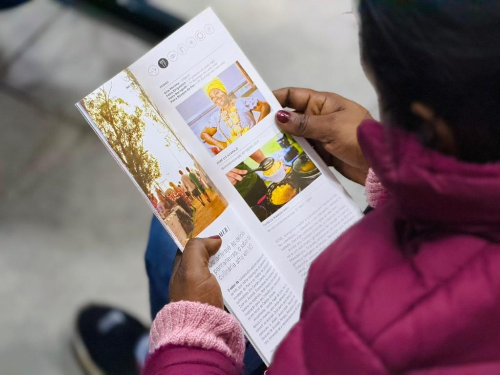 Visão de cima de uma pessoa lendo o guia, que mostra fotos de culinária típica, como o acarajé, e textos informativos sobre a cultura afro em MS.