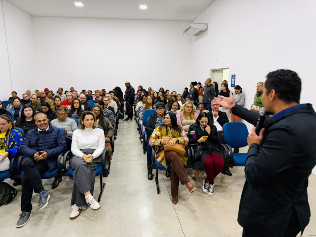 Ampla visão de um auditório com diversas pessoas sentadas, ouvindo um palestrante de terno que gesticula à direita da imagem.