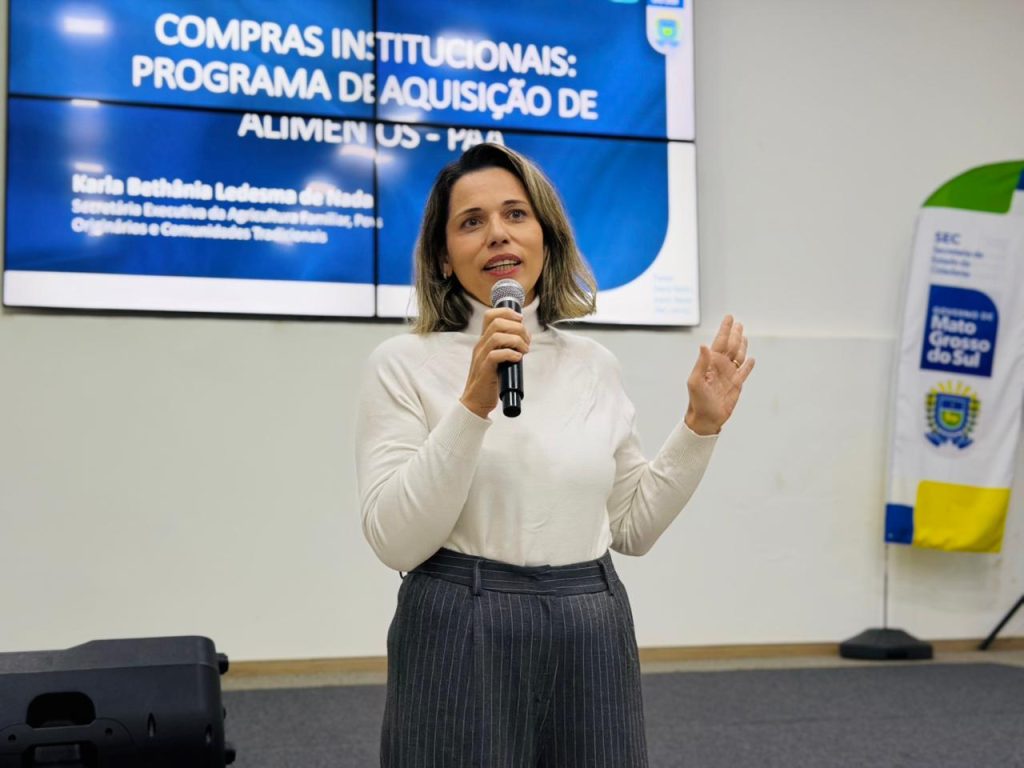Fotografia colorida em plano médio de uma mulher branca, com cabelos castanhos na altura dos ombros, falando ao microfone. Ela veste uma blusa branca de gola alta e calça social cinza risca-de-giz. Sua mão esquerda está levemente erguida enquanto ela discursa. Ao fundo, um telão azul exibe o texto: "COMPRAS INSTITUCIONAIS: PROGRAMA DE AQUISIÇÃO DE ALIMENTOS - PAA". À direita, vê-se um banner com o brasão do Governo do Estado de Mato Grosso do Sul.